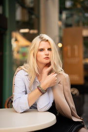 Woman sitting at a table outdoors with a blurred background. She wears 3 of the Mini Collection, diamond and gemstone bespoke bracelets by Caravan  of Stones. 