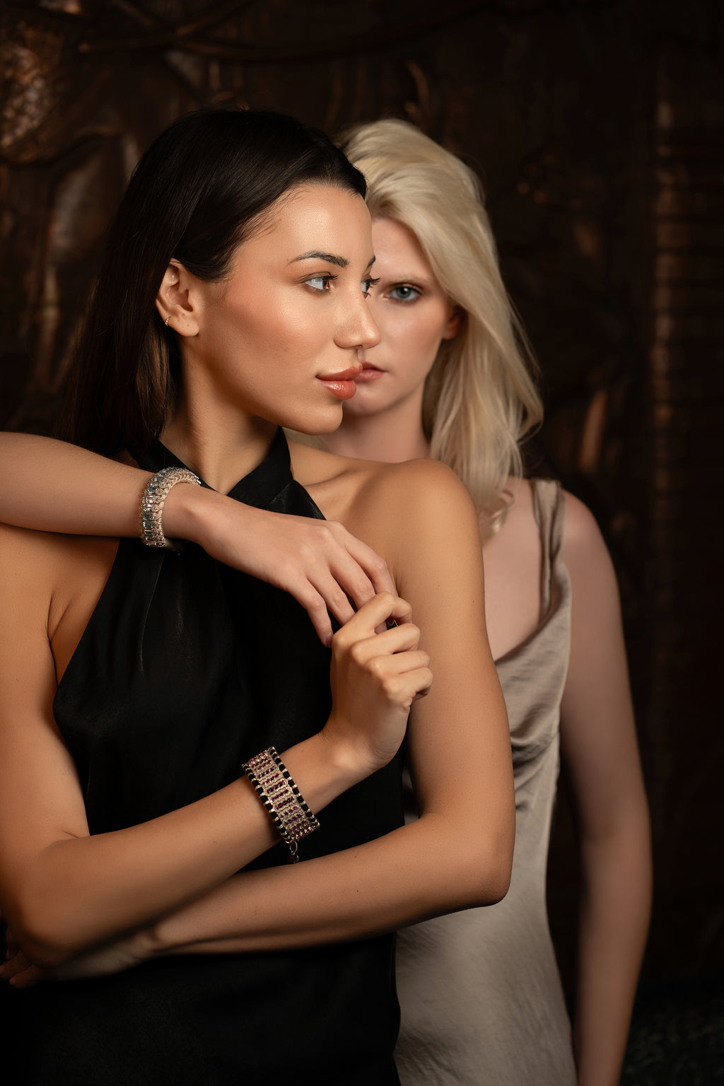 Two women standing close together with a dark background. The both wear stunning bespoke diamond and gemstone bracelets by London Brand, Caravan of Stones. 